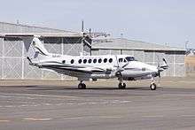 Colour photograph of a white aircraft on the tarmac of an airport in front of a grey building