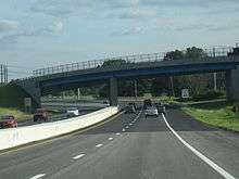 Ground-level view of a divided four-lane freeway; directly in front a large, curved bridge crosses over the freeway.