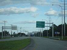 Ground-level view of a four-lane road; a green directional sign is posted above the route, and a traffic signal is visible in the distance.