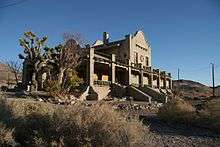 An ornate, two-story masonry building rests under a cloudless blue sky in a setting of gravel, two spiny trees, and many low shrubs. Its windows are boarded.