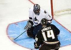 An ice hockey goaltender wearing a white jersey on his knees to make a save. He is looking downwards to the right as an opposing player in black skates towards him.