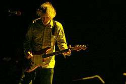 A man performing live on-stage, playing a red guitar. A yellow light shines on him.