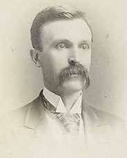 A dark-haired man in his early forties with a long, black mustache. He is wearing a black coat, white shirt, and black tie.