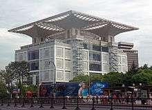 A four-story smooth-surfaced modern white building rising above trees and a street in the foreground with a spreading, partially latticed