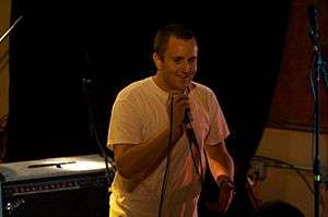 A man in a white T-shirt smiles and grasps a corded mic at its top