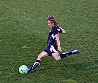 A white woman wearing a black shirt and black shorts while kicking a football.