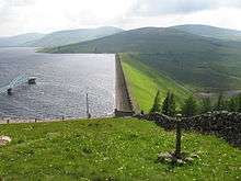 Daer Reservoir and dam with a blue footbridge extending out into the water and mown grass banks surrounding and a stone wall leading up to the dam