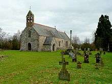 A stone church seen from the southwest, with a slate roof and red tiled ridge. At the west end is a door a window and a double bellcote; protruding from the south side is a porch