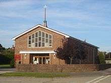 Three-quarter view of a simple brick church behind a low, curved wall. A flat-roofed entrance porch sits below a large, concrete-dressed, pentagonal, seven-light window.  On the roof just above this window is an extremely thin spire on top of a greenish metal ball, topped with a bird-shaped device.