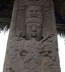 Close up of upper portion of an intricately carved stela, showing the face of a king with elaborate headdress and jewellery