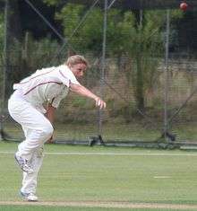 A woman with light brown hair wearing white cricket uniform with red piping has just bowled a cricket ball, still visible at the top right. She is bent over with her right arm extended in front of her.  Cricket training nets are visible in the background.