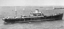 A dark-coloured merchant ship at sea with land and several other ships visible in the background