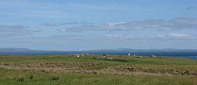 View looking north showing grass fields in the foreground, with ruined buildings visible in the middle distance and sea and islands visible on the horizon