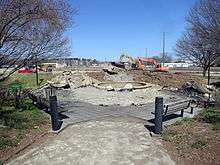 Construction equipment has begun to dig up concrete and level the earth beyond an area with a concrete fountain and wooden benches