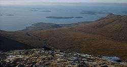 A view from a high rocky eminence with brown moorland below and a vista of small brown islands scattered in the sea beyond. A low bank of fog obscures the horizon.