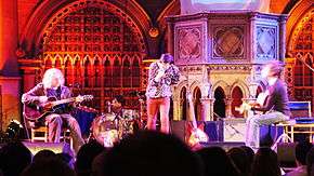 Three men performing in a renovate chapel. The man on the left plays an acoustic guitar, the man in the middle sings and the man on the right plays an acoustic bass guitar.