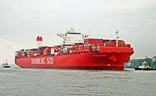 A large red ship, moving parallel to the shoreline, accompanied by two small tug-boats