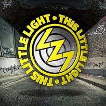 Image shows the acronym "LZ7" in a yellow font in the style of a lightning bolt on top of three concentric grey circles. The grey circles are enclosed within a thin yellow circle, which is in turn enclosed within a circle of the words "THIS LITTLE LIGHT·THIS LITTLE LIGHT·" in a yellow font. This entire logo is set against a background of a black-and-white perspective view of an urban street at night, showing a road, walls and dark sky. The walls have graffiti on them.