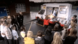 A white ice cream van is parked on a car park in front of a building. A man in a visor and a white jacket is hanging out of a side window of the van, and is handing a glowing object to a young girl in a red jumper and black skirt. Twenty-three similarly-aged children stand in a horseshoe shape around the girl.
