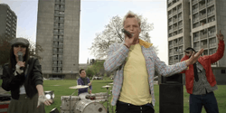 A young woman in a dark leather jacket, a man in a blue jumper, a man with bleached blonde hair in a yellow T-shirt and a man in a red leather jacket are performing on a grassy area outside a tower block estate during daytime. The man in the blue jumper is drumming at a drum kit; the other three are singing into microphones.