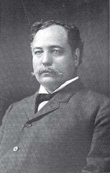 A portly man with black, wavy, receding hair wearing a black jacket and tie and white shirt