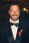 A bearded man, wearing a white shirt, black bowtie, and black jacket, smiles at the camera.