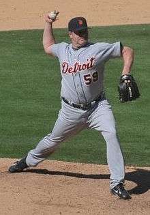 A man wearing a gray baseball uniform and dark baseball cap throwing a baseball with his right hand