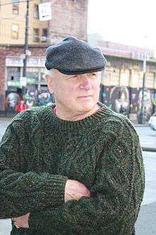 a pale elderly man in a sweater and flat cap