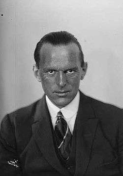 A head and upper torso portrait of Tryggve Gran, middle-aged man with receding hair and a weathered complexion, looking directly to camera and wearing a suit and tie