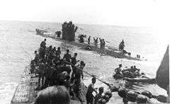 Three-quarter front view over the bow from a submarine conning tower of another submarine with numerous people standing on both submarines, at sea.