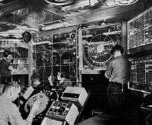 Men stand at various terminals scattered around a cramped room