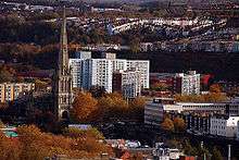 A view looking down towards a tall church spire which rises high above the surrounding trees and large modern buildings, with rows of hillside housing in the distance