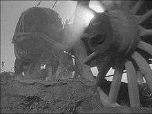 A wooden wagon wheel with 16 spokes occupies the right half of the image; the camera was perhaps 3 or 4 feet from this wheel. The wheel is biting about 6 inches into loose soil. Rays of sunlight are coming through just a few of the upper spokes, and are readily visible in the dusty air. The left half of the image shows the back side of a horse in harness that is straining to pull the wagon.