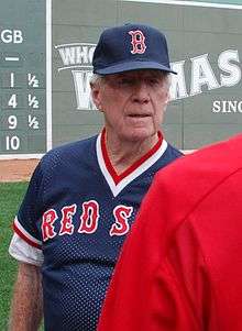 A white-haired man wearing a navy blue baseball jersey and cap