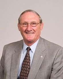 A portrait shot of William Suter, looking straight ahead. He has short gray hair and is wearing a light gray blazer with a maroon patterned tie over a light blue collared shirt. He has an American flag pin on his lapel.